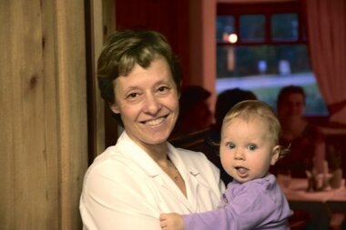 A woman smiles next to a wall while holding a small child in her arms.