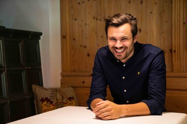A man with short hair sitting at a table in a wooden room, smiling.