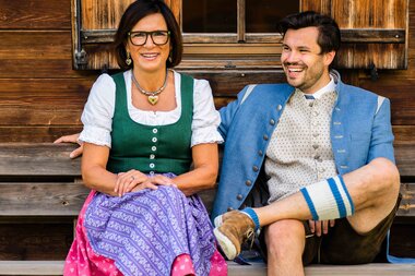 A couple in traditional Bavarian attire smiles while sitting on a wooden bench in front of a wooden wall.