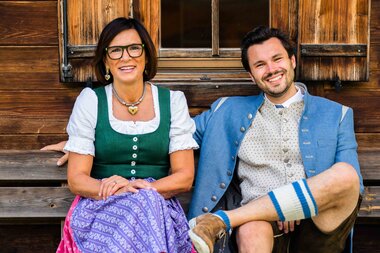 Ein Mann und eine Frau in traditioneller Tracht sitzen auf einer Bank vor einem Holzgebäude.