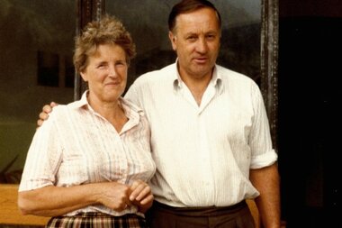 Ein Mann und eine Frau stehen vor einem Gebäude und lächeln. Die Frau trägt einen karierten Rock und eine Bluse, der Mann ein helles Hemd und braune Hosen.