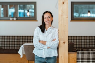 A smiling woman in a white shirt stands with crossed arms next to a wooden pillar in a modern café interior.