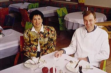 Ein Mann im Kochkostüm und eine Frau in blühender Bluse sitzen an einem gedeckten Tisch in einem Restaurant.