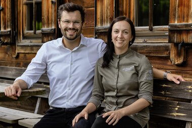 Zwei Personen sitzen lächelnd auf einer Bank neben einer Holzfassade. Der Mann in einem weißen Hemd und die Frau in einem grauen Koch-Outfit.