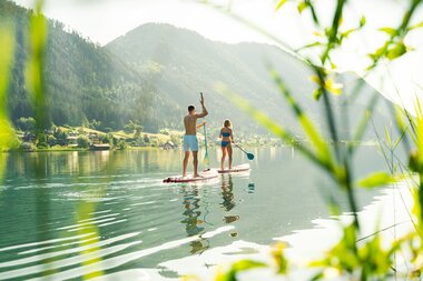 Ein Mann und eine Frau stehen auf Paddleboards auf einem ruhigen See, umgeben von Bergen, mit grünem Vordergrund.