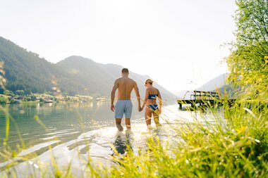 A couple walking hand in hand in shallow water by a lake, surrounded by mountains and greenery.