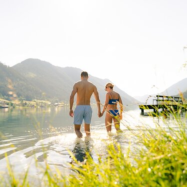 Ein Paar, das Hand in Hand im flachen Wasser eines Sees geht, umgeben von Bergen und Grün.