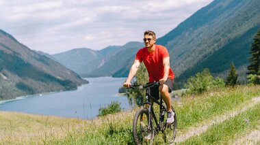 Ein Mann fährt mit dem Fahrrad auf einem grasbewachsenen Weg mit Blick auf einen See und umliegende Berge.