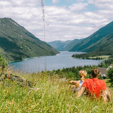 Ein Paar sitzt in der grünen Landschaft und schaut auf einen See und Berge.