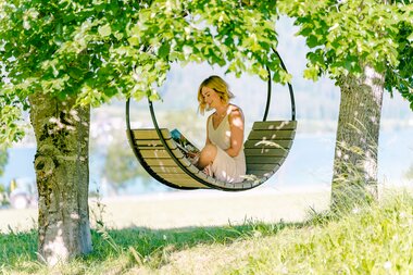 A woman in a white dress sits in a hanging swing between trees, reading a book.