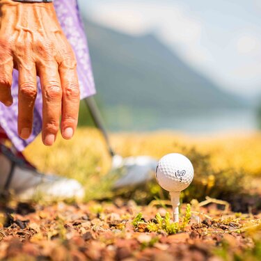A person in traditional clothing prepares to swing at a golf ball on a tee, surrounded by nature.
