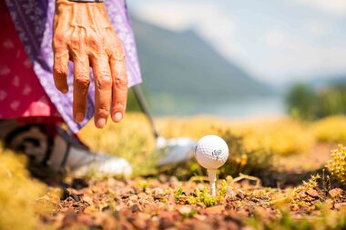 A person in traditional clothing prepares to swing at a golf ball on a tee, surrounded by nature.