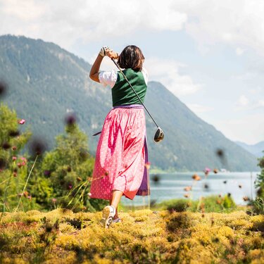 Eine Frau in traditioneller Tracht schwingt einen Golfschläger mit malerischen Bergen und einem See im Hintergrund.