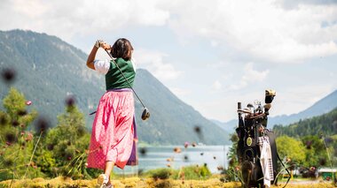 Eine Frau in traditioneller Tracht schwingt einen Golfschläger mit malerischen Bergen und einem See im Hintergrund.