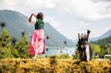 A woman in traditional attire swings a golf club with scenic mountains and a lake in the background.