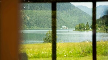 Blick auf einen ruhigen See, umrahmt von Fensterrahmen, umgeben von üppigen grünen Bergen und Bäumen.