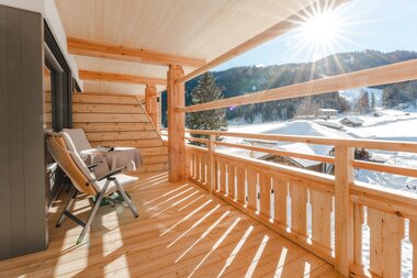 Sunlit balcony with wooden railing and seating, overlooking a snowy landscape and mountains.