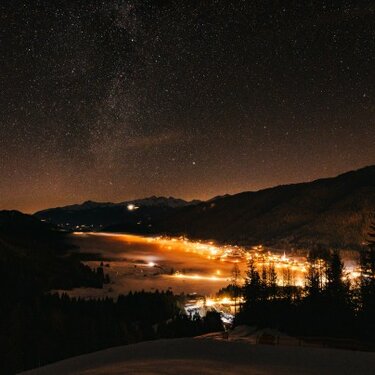 A starry night over a mountain valley with a glowing village near a lake.