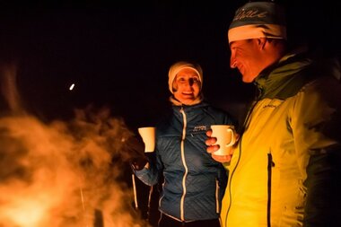 Two people in warm winter clothing stand by a campfire, holding cups.