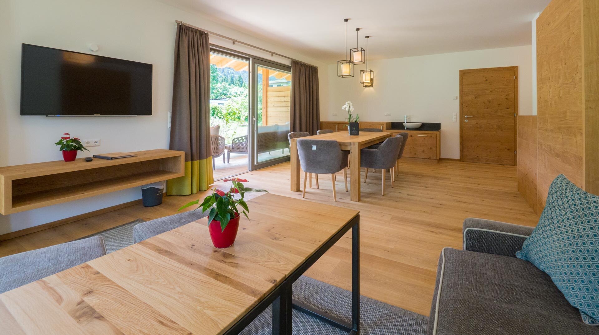 Helles, modernes Wohnzimmer mit Holzmöbeln, Fernseher und offener Essküche, Zugang zur Terrasse.
