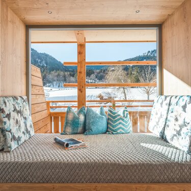 Gemütlicher Innenbereich mit weichen Kissen, der durch ein großes Fenster auf eine verschneite Landschaft blickt.