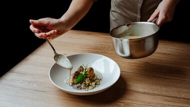 Eine Person gibt mit einem Löffel Soße über ein feines Gericht in einem weißen Teller.