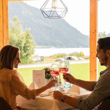 Ein Paar prostet sich mit Gläsern rosa Getränke an einem Tisch mit Blick auf einen malerischen See zu.