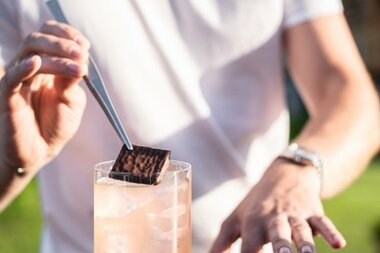 Ein Barkeeper platziert ein Stück Schokolade auf einem Getränk mit Eis in einem klaren Glas.