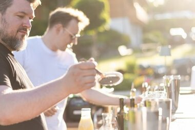Two men mixing cocktails at an outdoor bar in sunlight.