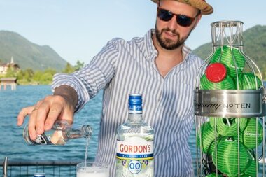 Bartender mit Sonnenbrille und Hut, der Gin in ein Glas mit Eis gießt, vor einer schönen Wasserlandschaft.