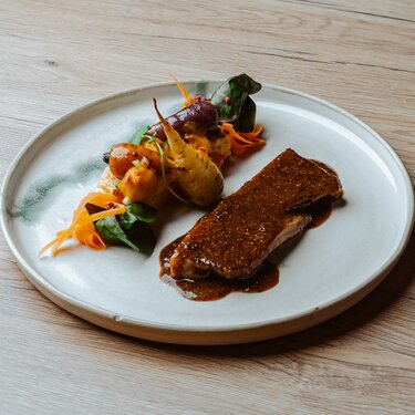 Ein kunstvoll angerichtetes Gericht mit einem Stück gegrilltem Fleisch in reicher Sauce, begleitet von buntem sautiertem Gemüse auf einem runden weißen Teller.