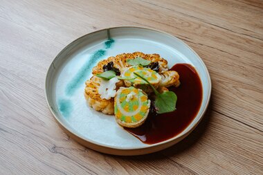 Kunstvoll angerichteter gerösteter Blumenkohl mit bunten Saucen und Garnierungen auf einem runden Teller.