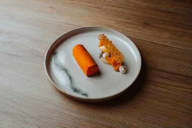 Ein angerichtetes Dessert mit einem orangen zylindrischen Kuchen neben einer dekorativen Anordnung aus gelben und weißen Elementen.