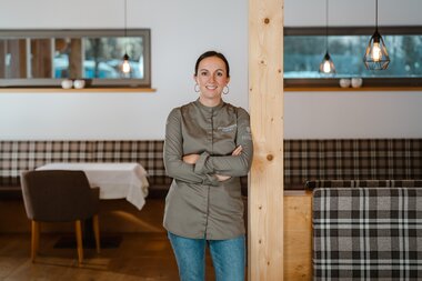 Eine Frau mit langen dunklen Haaren, die eine grüne Kochjacke trägt, steht selbstbewusst mit verschränkten Armen in einem gemütlichen Restaurant.