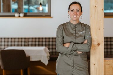 Eine lächelnde Köchin mit einem grauen Chefjackett steht an einer Säule in einem Restaurant.