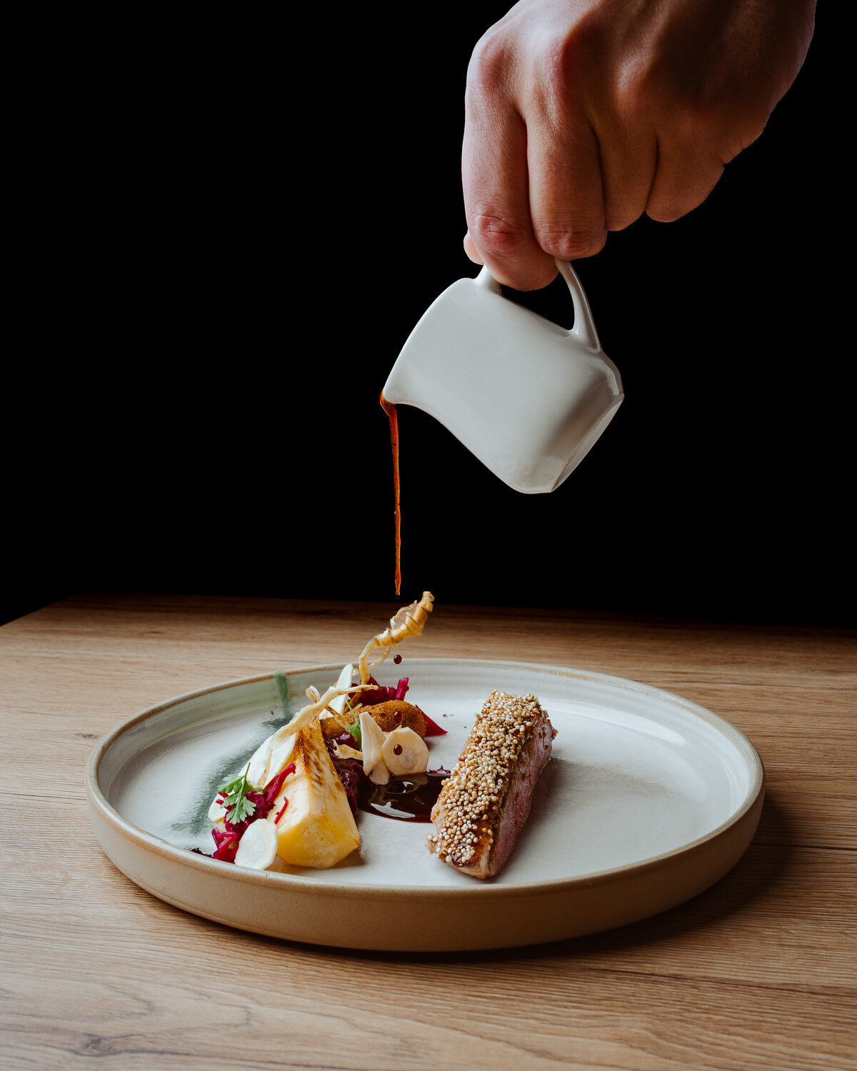 Eine Hand gießt Sauce aus einem kleinen weißen Kännchen über einen elegant angerichteten Gourmetteller.