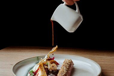Eine Hand gießt Sauce aus einer kleinen Kanne über einen kunstvoll angerichteten Teller mit Fleisch, Gemüse und Garnituren.