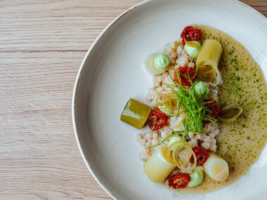 Ein kunstvoll angerichtetes Gericht mit Getreide, Lauch, getrockneten Tomaten und grüner Sauce.