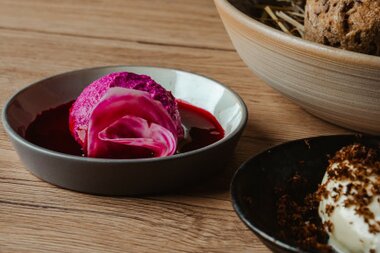 Ein Stück leuchtend pinkes Dessert auf einem weißen Teller, begleitet von roter Soße, mit zwei weiteren Schüsseln im Hintergrund.