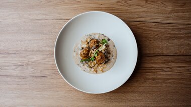 Ein kunstvoll angerichtetes Gericht auf einem weißen Teller, bestehend aus drei runden, gebratenen Klößen, umgeben von einer Sahnesauce und dekoriert mit feinen Kräutern.