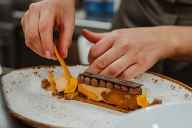 Die Hände eines Kochs dekorieren vorsichtig ein Dessert auf einem weißen Teller, das Schokolade und Orangenbestandteile enthält.