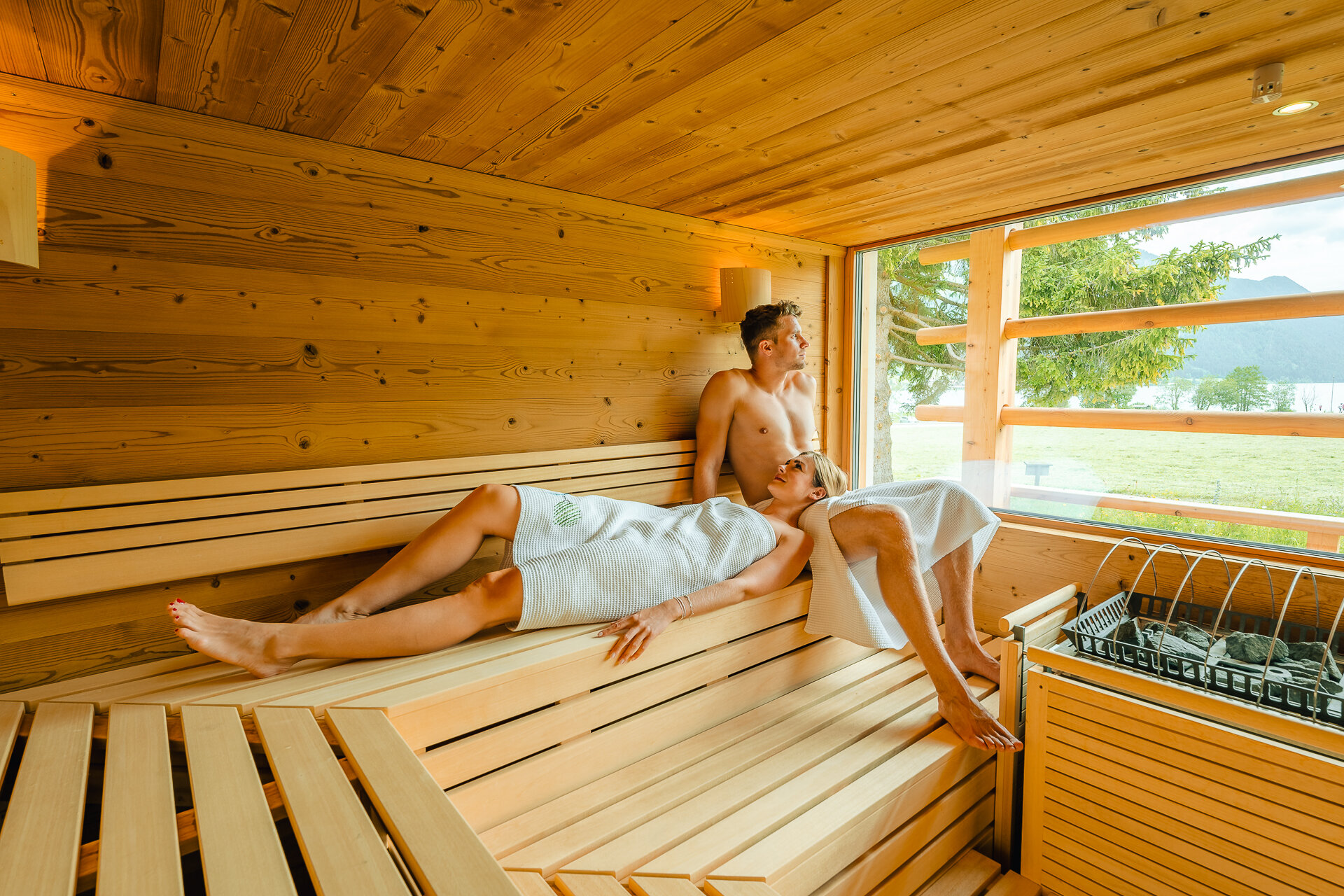 Zwei Personen entspannen in einer Holzsauna, eine sitzt auf der Bank, die andere liegt. Fenster mit Aussicht auf die Natur.