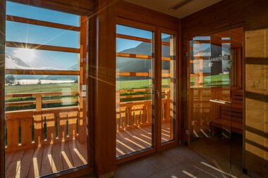 View of a sauna with large windows overlooking sunny mountains and a lake.