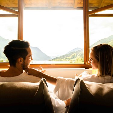 A couple in bathrobes seated by a large window with a mountain view, enjoying a cozy moment together.
