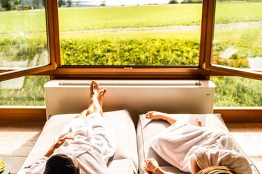 Zwei Personen in Bademänteln entspannen auf Sofas mit Blick auf einen malerischen See und Berge durch ein großes Fenster.