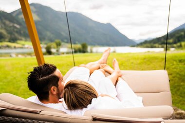 A couple in white robes relaxes together on a swing in a scenic outdoor setting, with mountains and a lake visible.