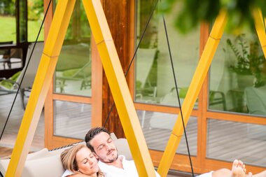 A couple relaxing in white bathrobes on a hammock, enjoying each other's company in a serene outdoor setting.