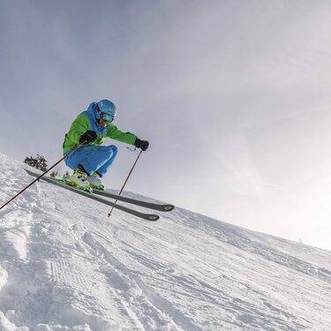 Ein Skifahrer in blau-grüner Kleidung springt über schneebedecktes Gelände vor einem hellen Himmel.