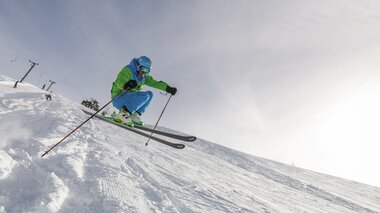 A skier in a blue and green outfit jumping over snow-covered terrain against a bright sky.