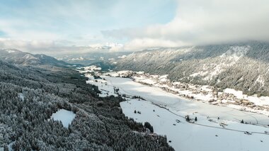 Luftaufnahme eines verschneiten Tals, umgeben von Bergen, mit einem kleinen Dorf in der Nähe des Flusses.